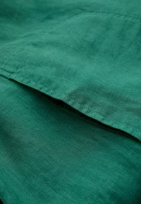 Vihreä pellavakankaan pinta, jossa on sileä rakenne ja hieno kiilto, näyttäen taitettuihin reunoihin ja yksinkertaisiin, siisteihin ompeluviivoihin.