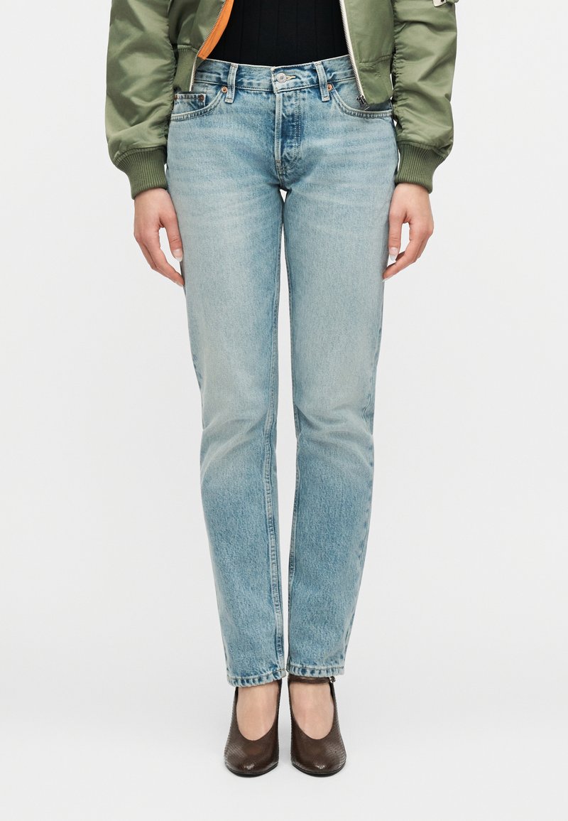 Άτομο που φοράει ανοιχτό μπλε τζιν παντελόνι με ίσια γραμμή, μαύρο μπλουζάκι, πράσινο μπουφάν τύπου bomber και σκουρό καφέ παπούτσια με τακούνι, στέκεται μπροστά σε απλό φόντο.