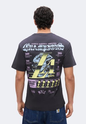 Άνδρας που φοράει μαύρο μπλουζάκι Los Angeles Lakers NBA Champions με εμφανή έτη και εικονίδια τροπαίων στην πλάτη, συνδυασμένο με μπλε τζιν.
