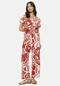 Rdeča in smetana cvetlična jumpsuit z kratkimi rokavi, pasom, široko nogo in lahkotnim materialom. Nosi se s sandali na vezi.