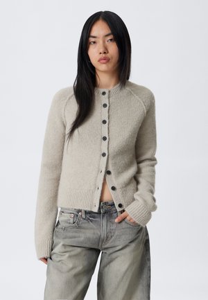 Young person with long black hair wearing a beige buttoned wool cardigan and loose gray denim jeans against a plain background.