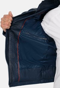 Giacca di pelle blu navy con un esterno liscio, dotata di una foderatura rossa brillante e una tasca interna con zip. Il tessuto sembra resistente e elegante.