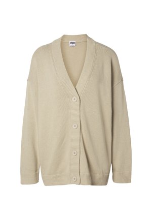 Femme debout, les mains dans les poches, portant un cardigan beige, un t-shirt blanc, un pantalon noir et des chaussures beiges sur un fond uni.