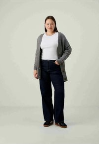 Cardigã cinza sobre uma camisa branca, emparelhado com jeans de perna larga azul escuro e sapatos pretos com sola castanha. Design simples com detalhes de botões.