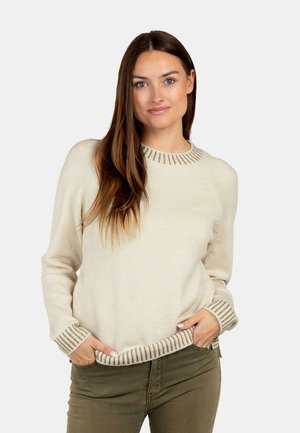 Jeune femme aux longs cheveux bruns portant un pull en maille crème avec des poignets et un ourlet à rayures, posant les mains dans les poches d'un pantalon vert olive.