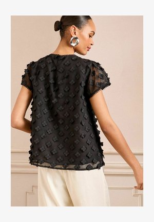 Schwarze Bluse aus transparentem Stoff, verziert mit strukturierten Blumenmustern. Sie hat kurze Puffärmel und einen gewellten Saum entlang der Kanten.