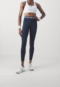 Granatowe legginsy Nike Pro z białym logo w pasie, połączone z białym sportowym biustonoszem Nike. Model nosi białe sneakersy z delikatnymi złotymi akcentami.