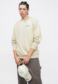 Νεαρός άνδρας με πλεξουδάκια φοράει μπέζ φούτερ Penfield και γκρι παντελόνι, κρατώντας λευκό καπέλο μπέιζμπολ μπροστά σε ένα απλό φόντο.