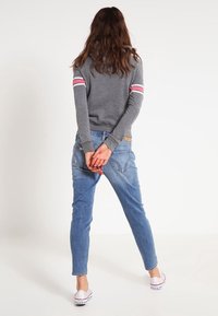 Suéter gris con rayas rosas y blancas en las mangas, combinado con jeans ajustados azul claro. La modelo está de pie con las manos entrelazadas detrás de la espalda.