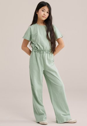 Mlada djevojka s dugom tamnom kosom nosi svijetlozeleno teksturirano kombinezon s kratkim rukavima, naboranim strukom i bijelim cipelama, stoji s rukama iza leđa.