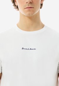 Weißes Baumwoll-T-Shirt mit navyblauem, gesticktem Text "Gassa d'Amante" auf der Brust. Kurze Ärmel und runder Ausschnitt.