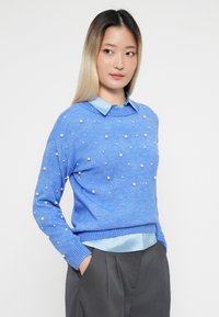 Jasnoniebieski sweter z dzianiny ozdobiony perłowymi detalami, okrągłym dekoltem i prążkowanymi mankietami, założony na jasnoniebieską koszulę z kołnierzykiem i szare spodnie.