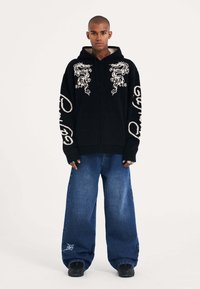 Felpa nera con zip e ricamo bianco a forma di drago, abbinata a jeans denim blu a gamba larga. Tessuto texture e vestibilità ampia.