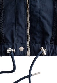 Close-up of navy jacket hem with metal zipper, snap button, mesh side panels, and adjustable drawstring cords with metal stoppers.