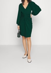 Femme portant une robe vert foncé à mi-longueur avec des manches longues bouffantes, tenant un sac à main vert assorti et portant des chaussures à talons blocs blanches.