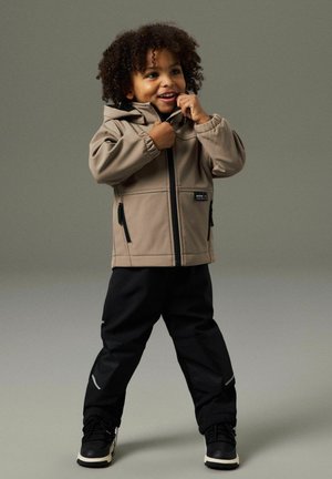 Dítě s kudrnatými vlasy v béžové bundě s kapucí a černých kalhotách, usmívající se a zapínající zip bundy.