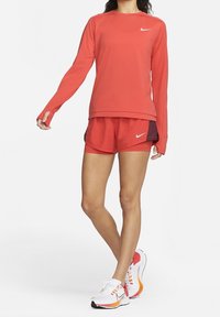 Mujer vestida con camiseta de manga larga roja de Nike y pantalones cortos a juego, con zapatillas de running Nike blancas y naranjas, de pie sobre un fondo blanco.