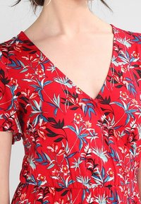 Czerwona sukienka w kwiaty z dekoltem w kształcie litery V i krótkimi rękawami, z niebieskimi i białymi wzorami kwiatowymi, teksturowanym materiałem i wciągniętą talią.
