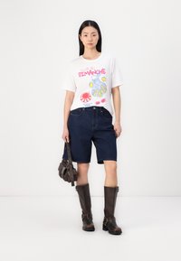 Hvit t-skjorte med fargerik grafisk trykk, blå denimshorts og brune knelangstøvler. Modellen holder en liten brun håndveske.