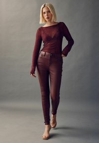 Burgundy långärmad topp med nätdetaljer, i kombination med figurnära burgundy läderbyxor och remsandaler i beige, mot en neutral bakgrund.