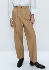 Bruna veckade byxor i slät tyg med hög midja och avsmalnande ben, i kombination med svarta ankellång stövlar. Vit skjorta med uppvikta ärmar.