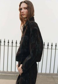 Jachetă din catifea neagră cu un design scurt, având închideri cu nasturi și două buzunare pe piept, plasată pe un fundal neutru.