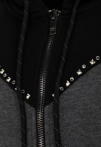 Schwarze und graue Kapuzenjacke mit Reißverschluss, verziert mit Nietenakzenten und Strasssteinen im oberen Bereich. Strukturierter Stoff mit Kordelzügen.