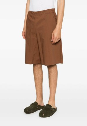 Uomo che indossa pantaloncini marroni larghi fino al ginocchio e scarpe slip-on verde scuro, in piedi davanti a uno sfondo semplice.