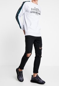 Homme portant un sweat-shirt gris clair avec du texte et une bande bleue sur les manches, un jean noir déchiré et des baskets noires, marchant avec les mains derrière le dos.