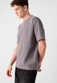 T-shirt en coton gris avec des manches courtes, col rond, détails de couture visibles et un logo subtil sur la poitrine. Texture douce et coupe décontractée.