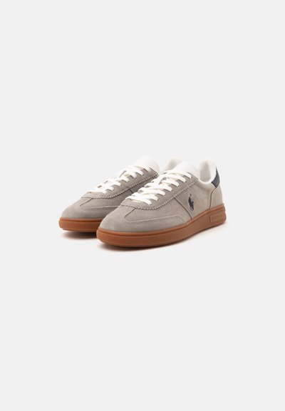 Pair of beige suede sneakers with white laces, brown rubber soles, dark blue heel detail, and a small dark horse logo on the side.