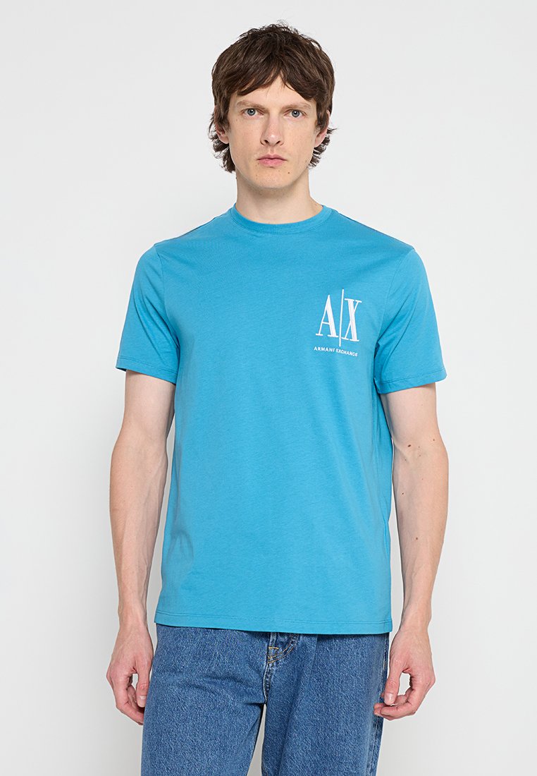 Młody mężczyzna w jasnoniebieskiej koszulce Armani Exchange z logo na piersi, w zestawieniu z niebieskimi dżinsami, stojący na jednolitym tle.