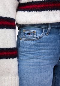 Κοντινή λήψη τσέπης μπλε τζιν με μικρό λογότυπο σημαίας και μέρος λευκού πλεκτού πουλόβερ με κόκκινες και ναυτικές ρίγες.