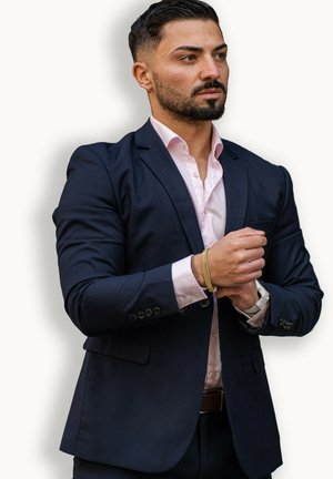 Marineblaues Sakko über einem hellrosa Hemd, mit einer Tasche und geknöpften Manschetten, dazu ein goldenes Armband und eine silberne Uhr am Handgelenk.
