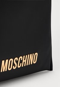 Must sõrk, millel on must sünteetilisest materjalist kott sileda tekstuuriga, millel on silmatorkav kuldne "Moschino" logo rasvases kirjas. Lihtne ristkülikukujuline disain.