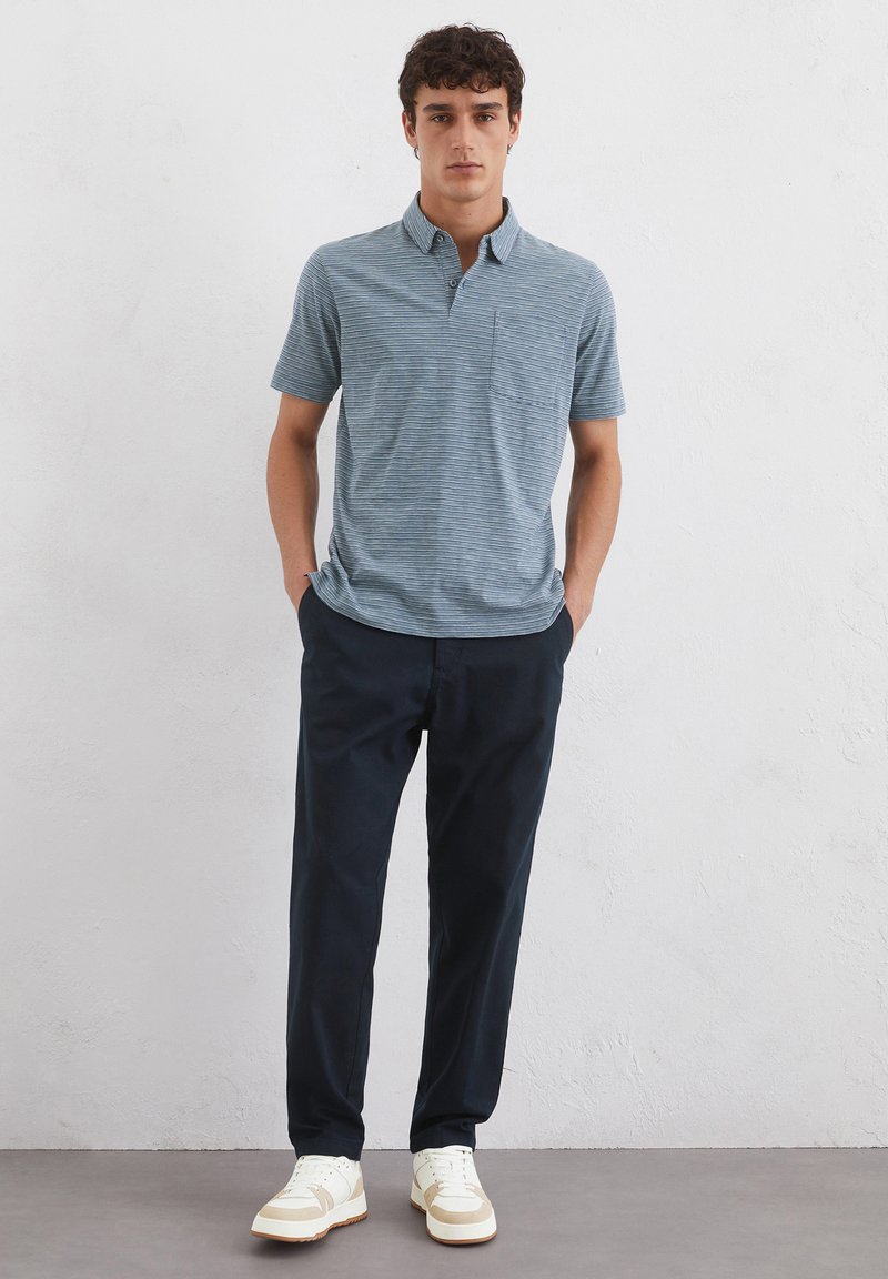 Kurzärmliges, blau gestreiftes Polohemd mit Kragen und Tasche, kombiniert mit dunkelblauen Hosen und weißen Sneakers mit hellen Akzenten.