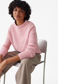 Maglione lavorato a maglia di un delicato rosa con colletto alto e texture a coste, abbinato a pantaloni beige, indossato da un modello seduto con disinvoltura.