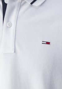 Polo blanca de tejido texturizado. Presenta una tapeta con dos botones y un pequeño logotipo bordado en rojo, blanco y azul en el pecho.