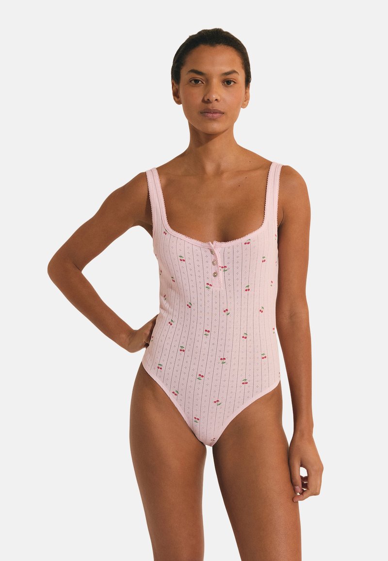 Femme debout avec la main sur la hanche, portant un body rose sans manches avec de petits imprimés de cerises et des détails de boutons à l'avant.