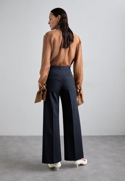 Brun ribbet bluse med bundet ærmer, parret med højt Midje, brede bukser i marineblå og hvide spidse hæle. Glatte teksturer og rene linjer.