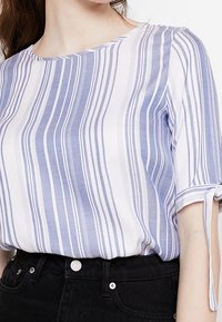 Lekka koszula w pionowe niebieskie i białe paski, okrągły dekolt i krótkie rękawy z wiązaniami. Noszona z czarnymi spodniami.