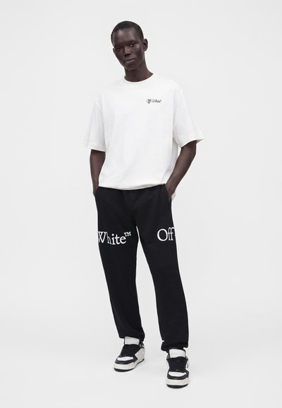 Άνδρας που φοράει λευκό μπλουζάκι Off-White και μαύρο παντελόνι με το κείμενο "Off White", στέκεται με τα χέρια στις τσέπες μπροστά σε απλό φόντο.