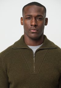 Brauner gerippter Strickpullover mit hohem Kragen und Reißverschluss. Über einem grauen T-Shirt getragen. Der Stoff erscheint dick und strukturiert.