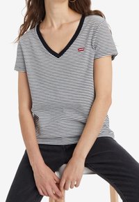 Osoba ubrana w czarno-białą w striped koszulkę V-neck z małym czerwonym logo Levi's, siedząca z rękami opartymi na kolanach.