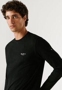 Mladi moški z ohlapnimi kodri, ki nosi črno majico z dolgimi rokavi Pepe Jeans London, stoji pred enostavnim svetlim ozadjem.