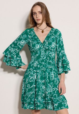 Jeune femme portant une robe verte à fleurs avec des manches cloche superposées et un ourlet volanté, posant devant un fond clair uni.