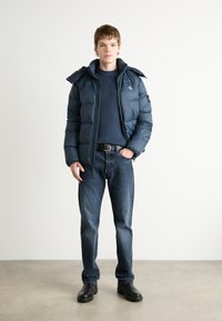 Donzen jas in donkerblauw, met een rits aan de voorkant, hoge kraag en zijzakken. Draagt over een ribbeltrui in marineblauw en gecombineerd met donkere jeans.