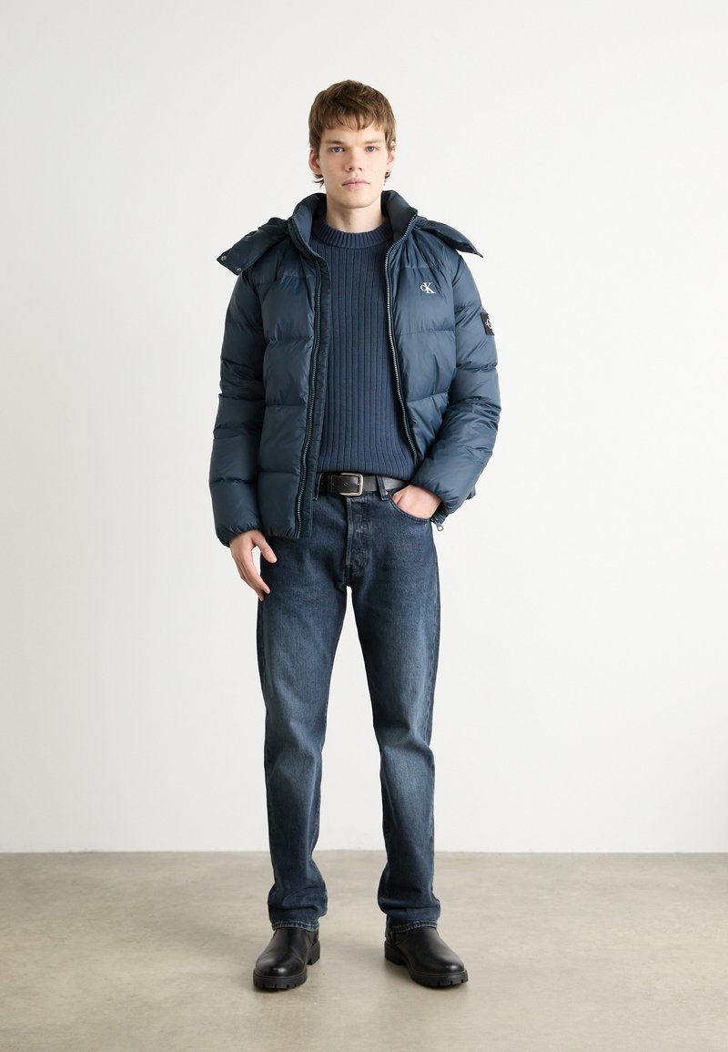 Dunkelblaue Pufferjacke mit Reißverschluss vorne, hohem Kragen und Seitentaschen. Getragen über einem gestreiften marineblauen Pullover und kombiniert mit dunklen Jeans.