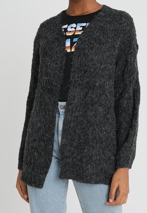 Cardigan gris foncé en matière pelucheuse, ouvert sur le devant, porté par-dessus un t-shirt noir à motif et un jean taille haute bleu clair.