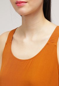 Top a maniche larghe color ruggine, realizzato in un tessuto liscio e strutturato, con un collo arrotondato che sottolinea la semplicità.